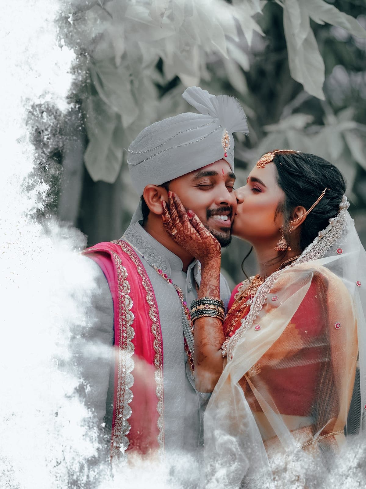 Bride in a lace veil kissing the groom on the cheek, surrounded by soft greenery