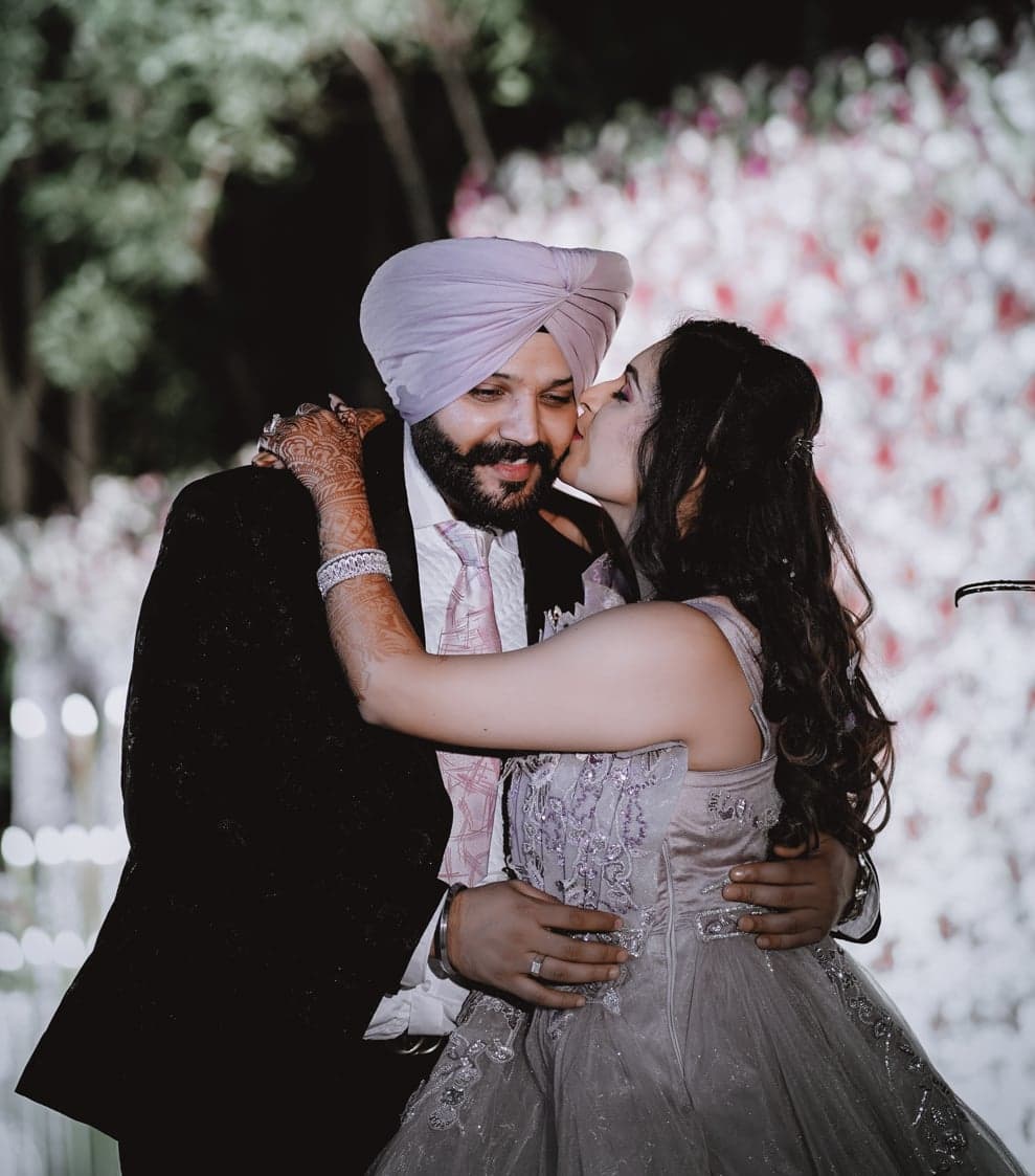 Bride in a shimmering lavender gown kissing her groom on the cheek in front of a flower wall at their reception